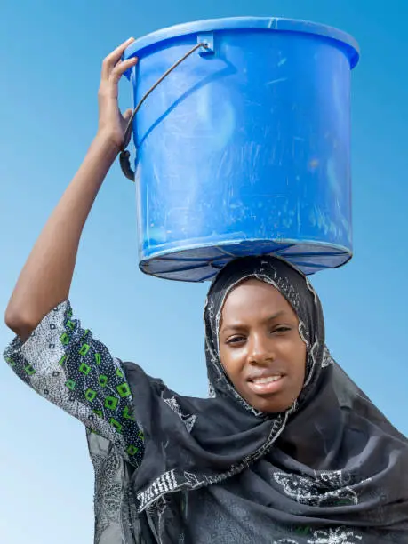 Young Afro beauty (eighteen years old) carrying a bucket of water on her head, photo