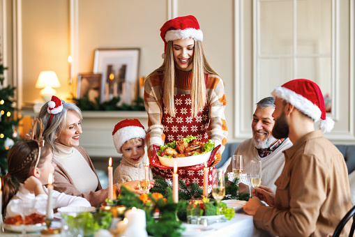Happy multi-generation family grandparents, parents and kids wearing Santa hats celebrating Christmas at home while sitting all together at table, young woman serving traditional New Years eve meal