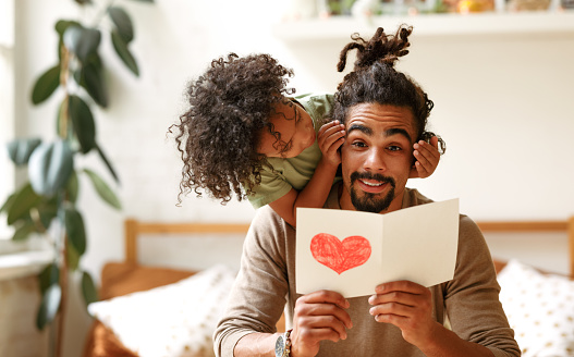 Surprise for daddy. Cute smiling afro american kid son giving father postcard with red drawn heart and covering his eyes with hands, happy child congratulating dad during family holiday at home