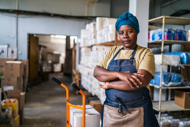istock Female african worker with working uniform and traditional headscarf,  pulling hand truck with boxes through  a warehouse 1357823650