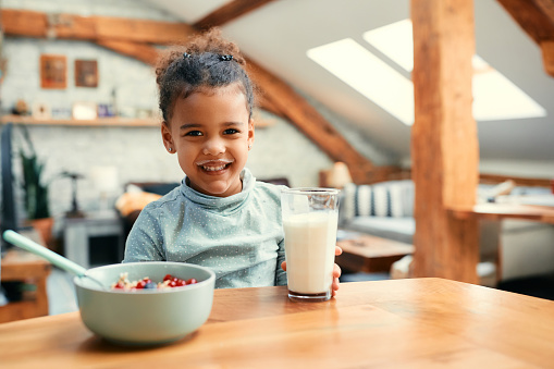 Happy African American little girl having milk mustache while eating breakfast at dining table and looking at camera.