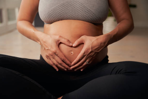 madre embarazada caucásica sentada en una esterilla de yoga formando una forma de corazón con las manos en el vientre - ewan fotografías e imágenes de stock