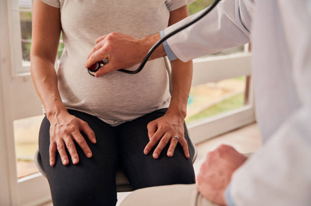 donna incinta caucasica seduta in clinica mentre il medico maschio esamina la pancia incinta con lo stetoscopio - ewan foto e immagini stock