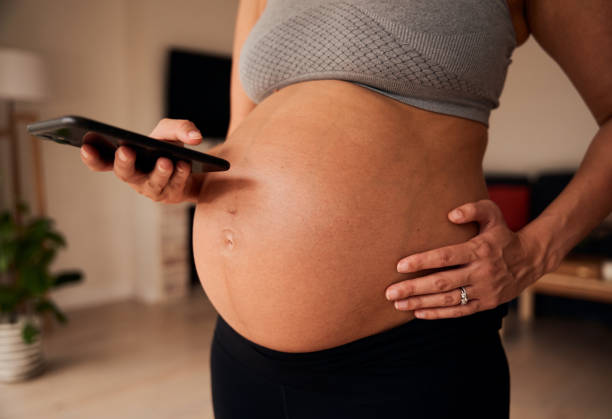 mujer embarazada caucásica de pie en la sala de estar enviando mensajes de texto en un dispositivo celular - ewan fotografías e imágenes de stock