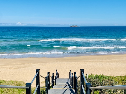 Horizontal seascape vanishing point of walking down stairs leading to turquoise breaking waves on empty beach with grass sand dunes under a clear blue sky day Australia