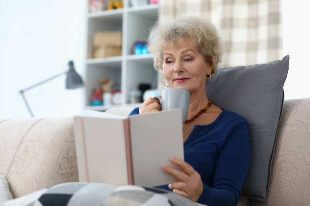 istock Old lady reading book and drinking cup of coffee being comfy on sofa at home 1357846259