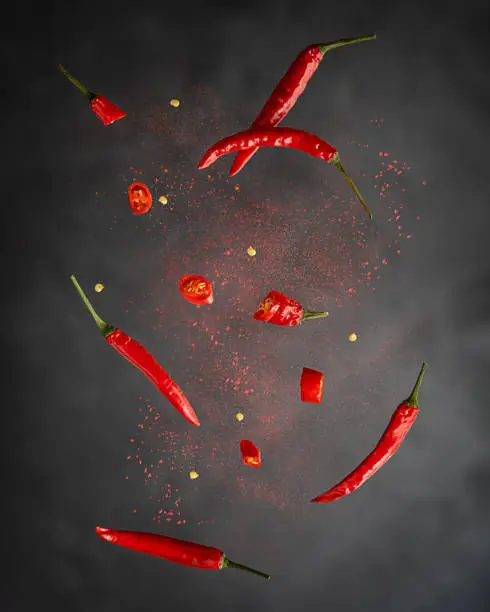Levitation or flying of red chili pepper whole and slices with paprika powder used for food seasoning to make hot spicy flavour in mexican and asian cuisine against dark black background. Vertical