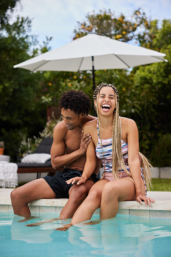 Young couple in swimwear laughing together while sitting at the edge of a pool with their legs in the water in summer