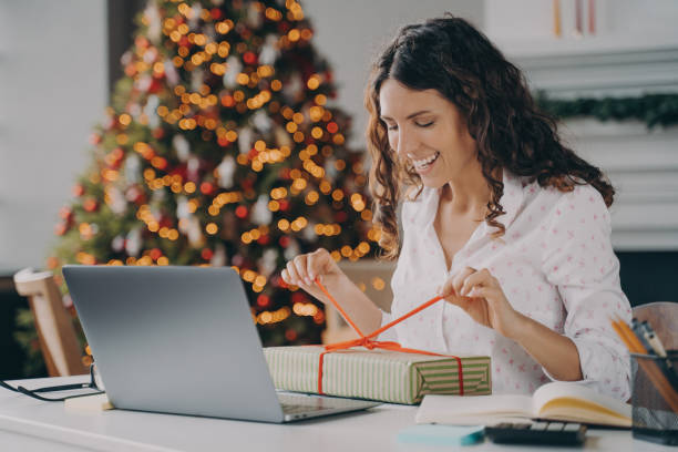 femme souriante déballant un cadeau de noël assis à un bureau avec un ordinateur portable contre un arbre de noël - christmas office gift people photos et images de collection