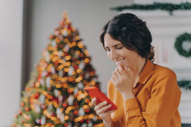 Overjoyed hispanic lady sits in living room decorated with Xmas tree looking at mobile screen