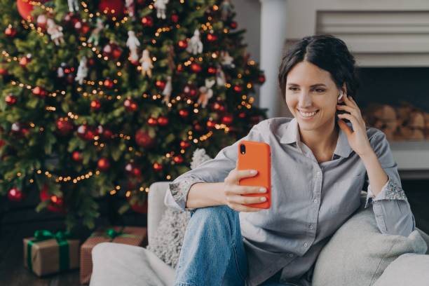 femme hispanique heureuse dans des écouteurs sans fil tenant le téléphone, assise près du sapin de noël à la maison - christmas tree audio photos et images de collection