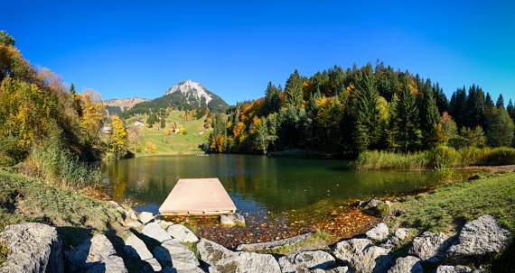 A beautiful pond with a colorful forest beside, a jetty in the foreground and mountains in the background during autumn. Vorarlberg, Fontanella