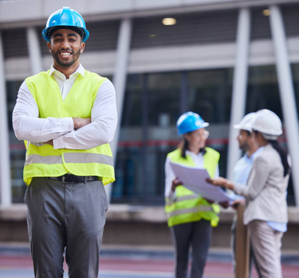istock Shot of a handsome young architect standing with his arms folded while his colleagues discuss behind him 1357867377