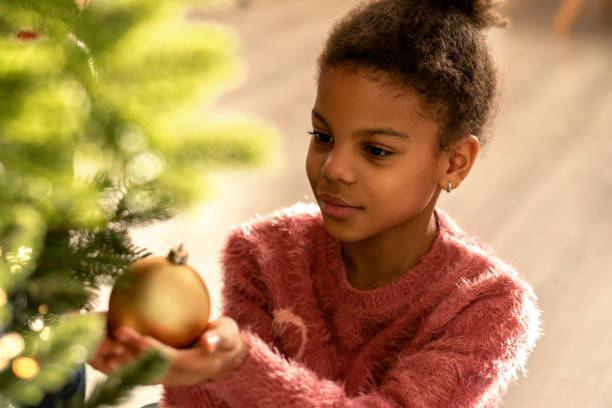 Nine-year-old African-American girl in a pink sweater is decorating Christmas tree at home,close up.New Year and Christmas concept.Diverse people.