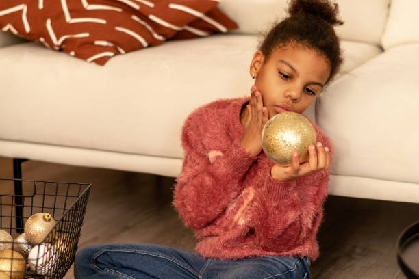 Nine-year-old African-American girl in a pink sweater looks at the golden Christmas ball at home.New Year and Christmas concept.Diverse people.