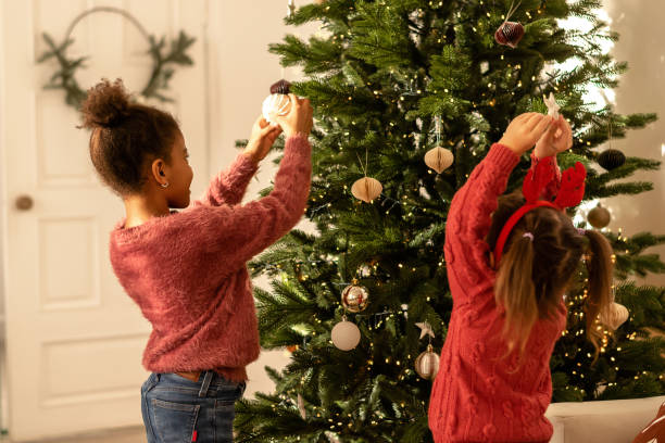 Two sisters in an interracial family decorate a Christmas tree at home.The girls are dressed in red sweaters and jeans.Rear view.New Year and Christmas concept.