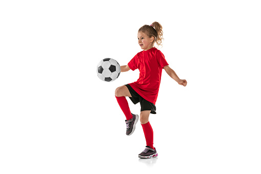 Portrait of girl, child, football player in red uniform training, kicking ball with knee isolated over white background. Concept of action, team sport game, energy, vitality. Copy space for ad.