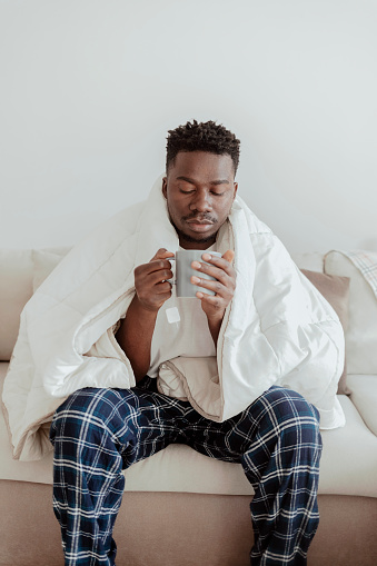 Shot of a young man having tea while recovering from an illness in bed at home. Sick African-American man drinking hot healing tea in bed, looking exhausted, empty space. Healthcare concept.