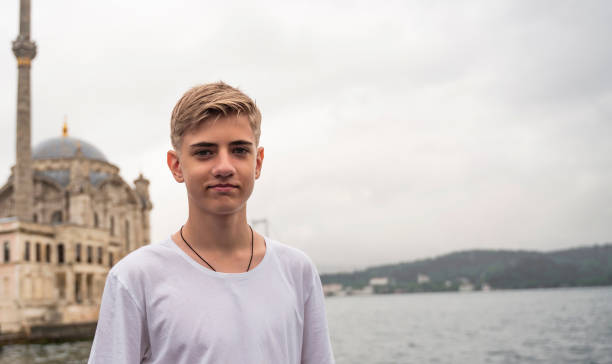 handsome young teenage man of 15-18 years old looks into the camera on a rainy day against the backdrop of the ortakoy mosque and the bosphorus bridge. - teenager 14 15 years 13 14 years cheerful imagens e fotografias de stock