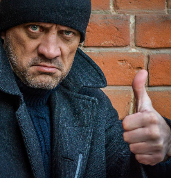 A man in a coat with a thumb up, portrait. A man with a confident and strong-willed look, against a background of a brick wall. Unshaven man in an autumn coat.