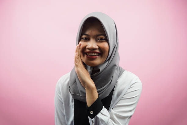 Beautiful young asian muslim woman smiling confidently and excitedly close to camera, whispering, telling secrets, speaking quietly, silent, isolated on pink background stock photo