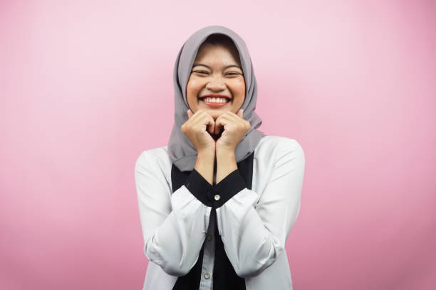 Beautiful young asian muslim woman smiling happy, cute, feeling comfortable, feeling cared for, feeling good, with hands holding chin isolated on pink background stock photo