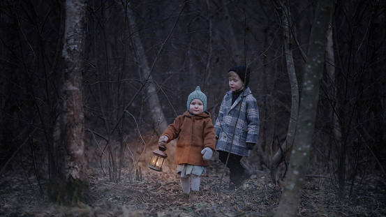 Small children, walk through the night scary autumn or winter forest. The girl carries a lamp with candle. They are lost and are looking for their parents. Fairy-tale scene escaping from monsters.