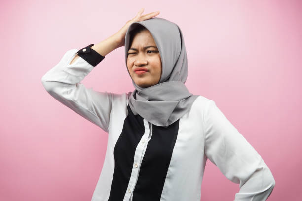 Beautiful young asian muslim woman stressed, dizzy, have a problem, feeling depressed, with hands holding head isolated on pink background stock photo