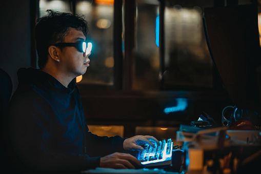 Asian Chinese male wearing glowing eyeglasses in front of desktop PC at night in low light dark room