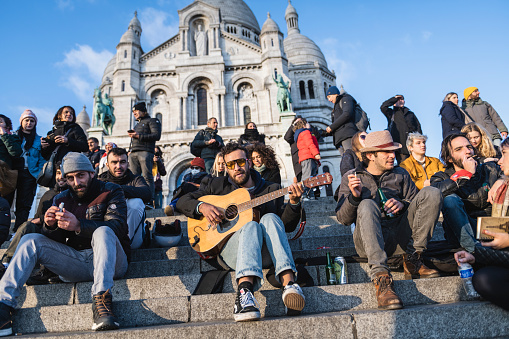 Paris, France - November 24, 2021: People from various countries sit on the steps in front of the Sacre-Coeur in Montmartre, one man singing as he plays the guitar.