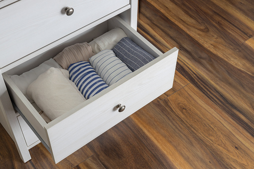 Top view of opened drawers of white dresser and rolled shirts in it.Empty wooden floor indoors