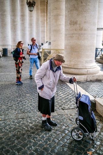 Vatican City, October 07 -- An elderly woman pushes her cart near a couple of tourists under Bernini's Colonnade, in the square of St. Peter's Basilica, in the historic center of Rome. Image in High Definition format.