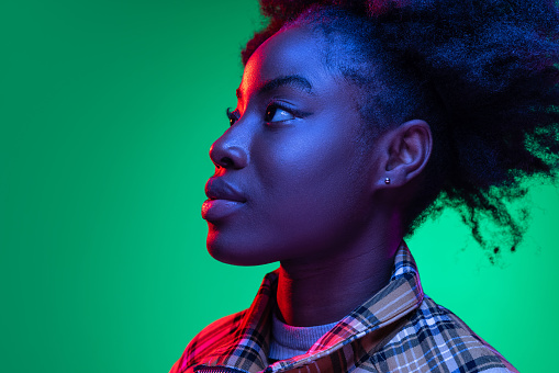 Looking away. Beautiful dark skinned young girl with adorable hairdo isolated on dark green studio background in purple neon light. Concept of human emotions, facial expression, youth, sales, ad.