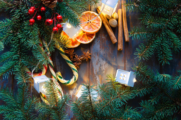 istock Christmas frame with fir branches, red berries, dry oranges, candy canes, lights and cons on dark blue wooden background. Christmas wallpaper or background. 1357898068