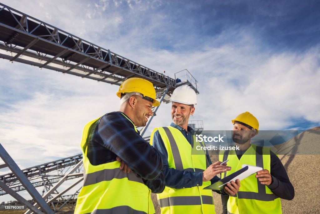 Open-pit mine workers Open-pit mine engineer and workers wearing protective clothes and helmets standing in front of pile of gravel, discussing plans. Adult Stock Photo