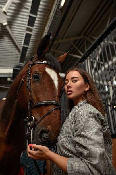 istock Smiling young woman standing inside a stable holding a saddle while preparing her chestnut horse for a ride 1357907335