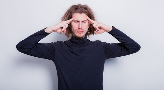 Closeup portrait man thinks very intensely having headache. Human emotions man in dark gray turtleneck and short dreadlocks on head touches his forehead with his fingers. Copy space grey background