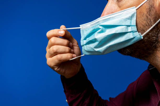 szczegół twarzy latynoskiego mężczyzny zdejmującego maskę chirurgiczną wyizolowaną na niebieskim tle, pozbywa się maski z rozpaczliwej sytuacji nowej normalności spowodowanej koronawirusem - protection mask zdjęcia i obrazy z banku zdjęć