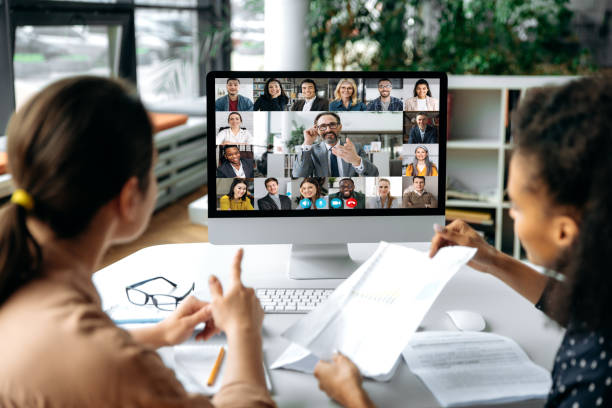 Virtual meeting online, video call. View over shoulders of two women to a computer screen with business leader and successful team, chatting by a video conference, discuss working issues, strategy stock photo