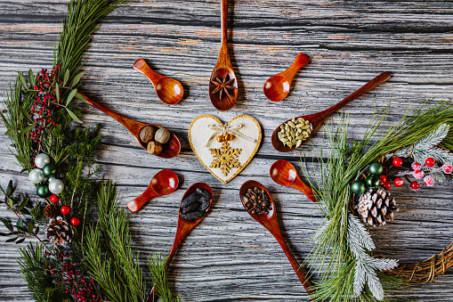 Beautiful flat lay of delicious Christmas spices in five dark wooden spoons and 4 empty spoons on rustic wooden grunge background arranged to a Christmas star with tonka bean, anise star, cardamom, clove, nutmeg and with christmas decoration on fir branch. Color editing with added grain. Part of a series.