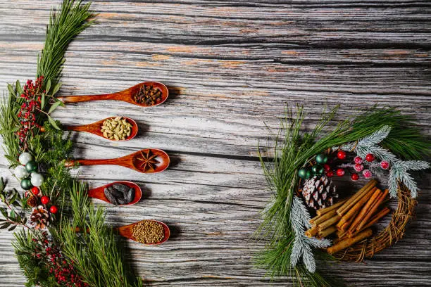 Flaylay of diferent Christmas spices in five dark wooden spoons on rustic wooden grunge background. The spices are tonka bean, cardamom, anise star, clove, cinnamon and coriander with christmas decoration on fir branch. Image composition is intended for cropping to banners. Copy space. Color editing with added grain. Part of a series.