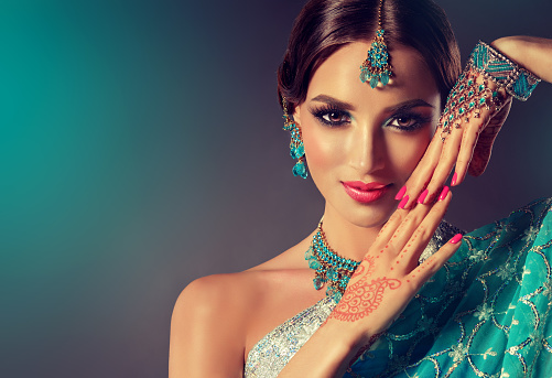 Young, brown haired woman with soft smile on the face is demonstrating perfect makeup with smokey eyeshadows. Fashionable jewelry items in Indian style is covers her forehead, ears, neck and palms with henna tattoo. Indian beauty.