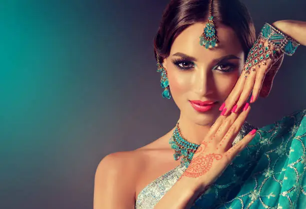 Photo of Young woman with soft smile on the face is demonstrating perfect makeup, jewelry in Indian style and  henna tattoo on the palms.