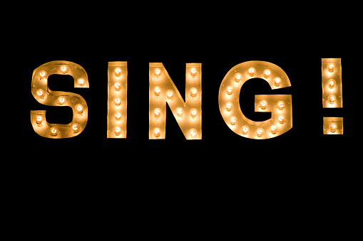 SING! lit sign, close-up on full frame black background with golden bright light bulbs of a style often used in theater and stage, performances.