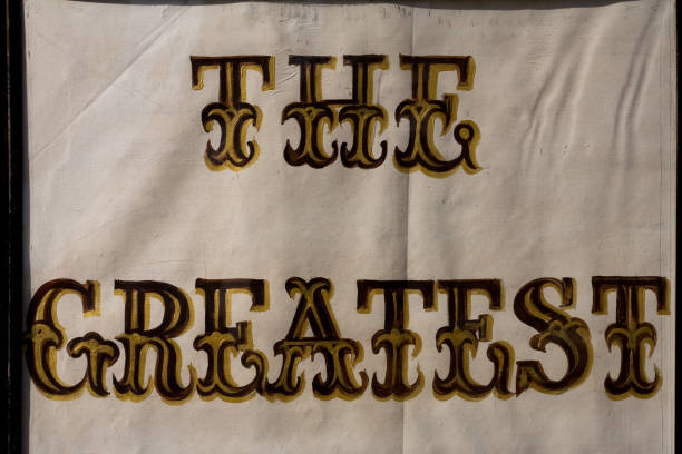 Hand painted window sign behind cracked display glass that the says THE GREATEST. Close-up and full frame, with black and gold decorative lettering on textured white paper