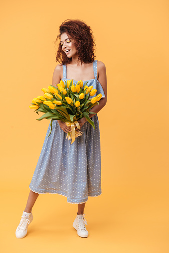 Full-length portrait of Smiling African woman in blue dress holding bouquet of flowers over yellow background