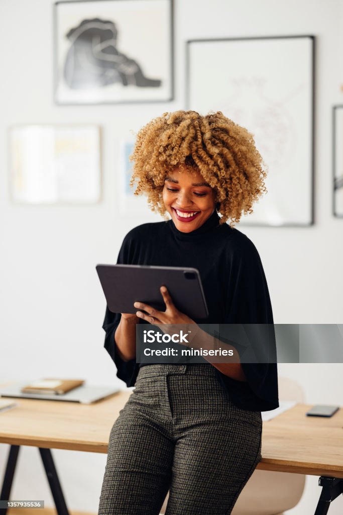 Happy Businesswoman Sitting on Her Desk Using Her Tablet Cheerful African American woman, sitting on her desk, smiling and using her laptop. She is typing something. Women Stock Photo