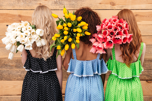 Back view of Three women in dresses standing  near the wall  and holding bouquets of flowers