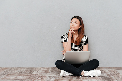 Young serious woman dreaming while sitting on floor and holding laptop isolated