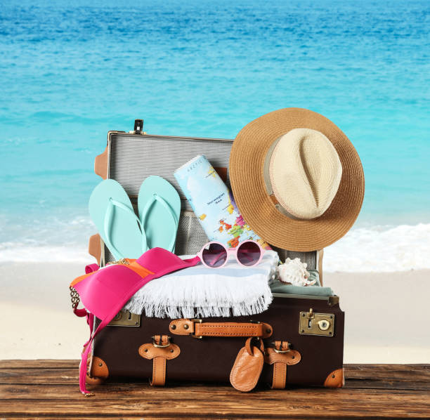 istock Open vintage suitcase packed for summer vacation on wooden surface against sandy beach 1357944929